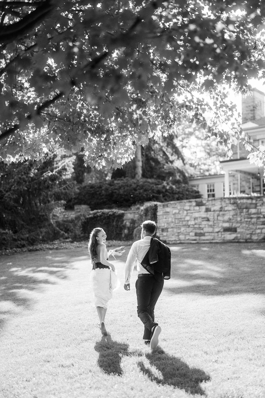 Anastasiia and Petr walk barefoot on grass under tree shade, stone wall and mansion behind them.