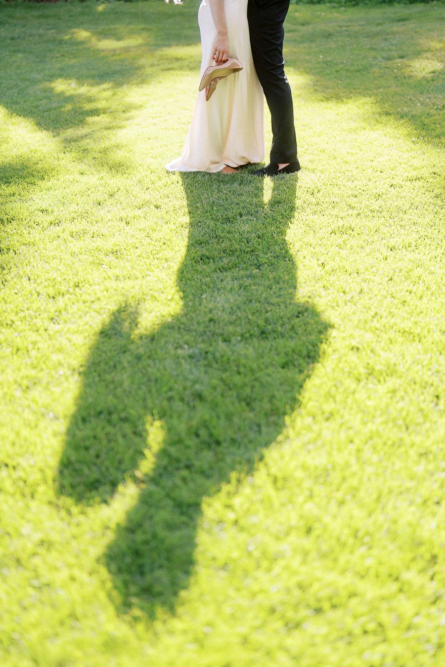 Engaged couple standing together on grass with long shadow and woman holding shoes.