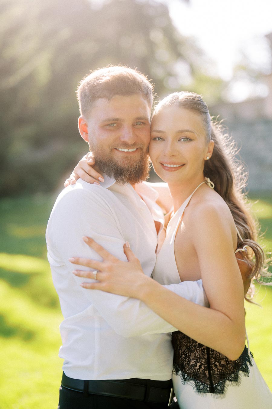Anastasiia and Petr embracing closely, smiling in bright sunlight with greenery behind them.