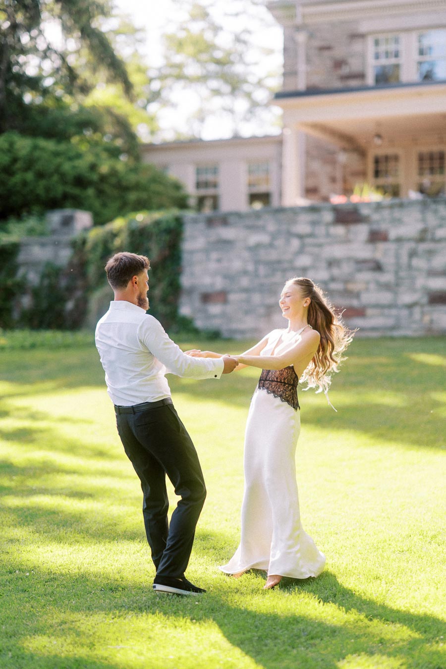 Engaged couple holding hands and spinning on a bright green lawn with stone mansion in background.