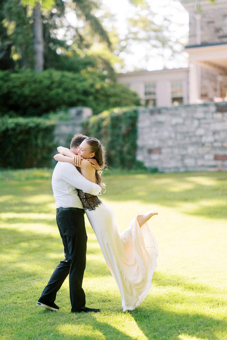 Engaged couple embracing as groom-to-be lifts bride-to-be on sunlit grass near stone wall.