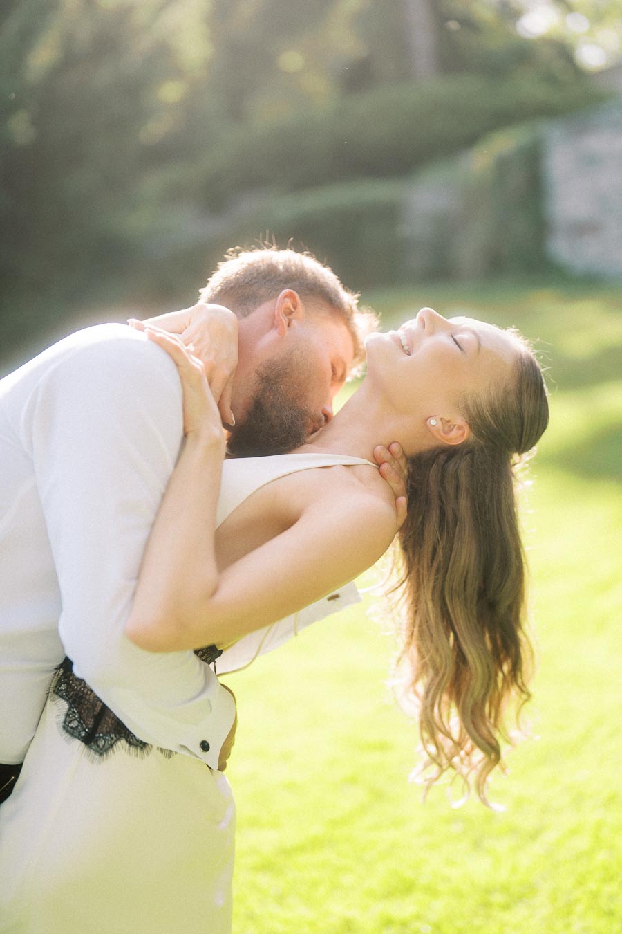 Engaged couple in white attire sharing a dip on sunlit grass with soft background.
