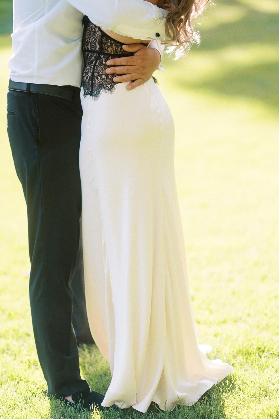 Engaged couple embracing closely on a bright green lawn, focusing on torsos and lower bodies.