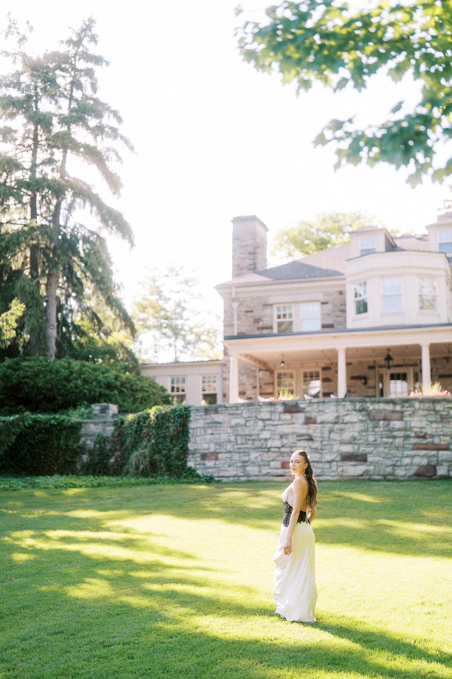 Woman in white dress stands on green lawn with Paletta Mansion and stone wall in background.