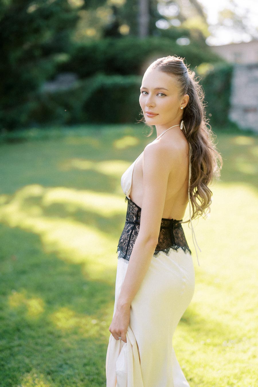 Anastasiia in a sleeveless light dress with black lace detail, standing on green grass in soft sunlight.