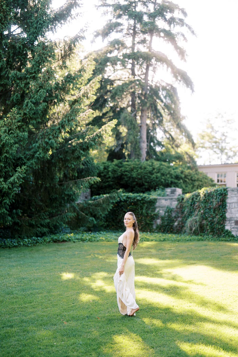 Anastasiia in a light dress standing on grass with trees and ivy-covered stone wall behind her.