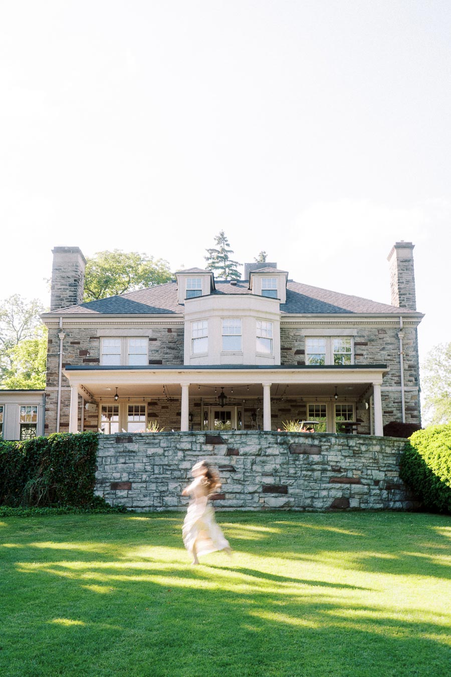 Woman in white dress spinning on green lawn in front of Paletta Mansion's stone facade.