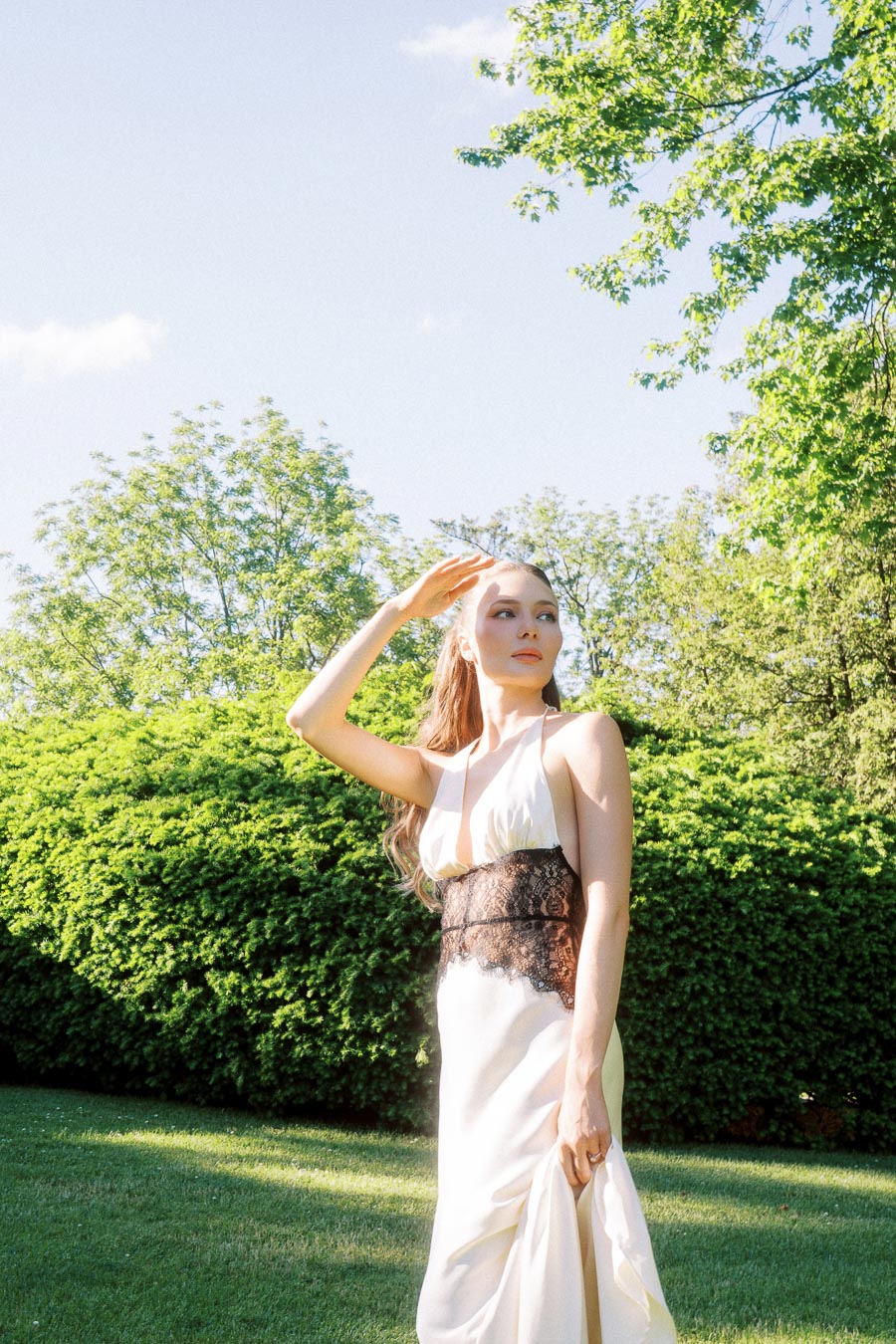 Anastasiia stands outdoors on a lawn, holding her dress and shading her eyes from sunlight.