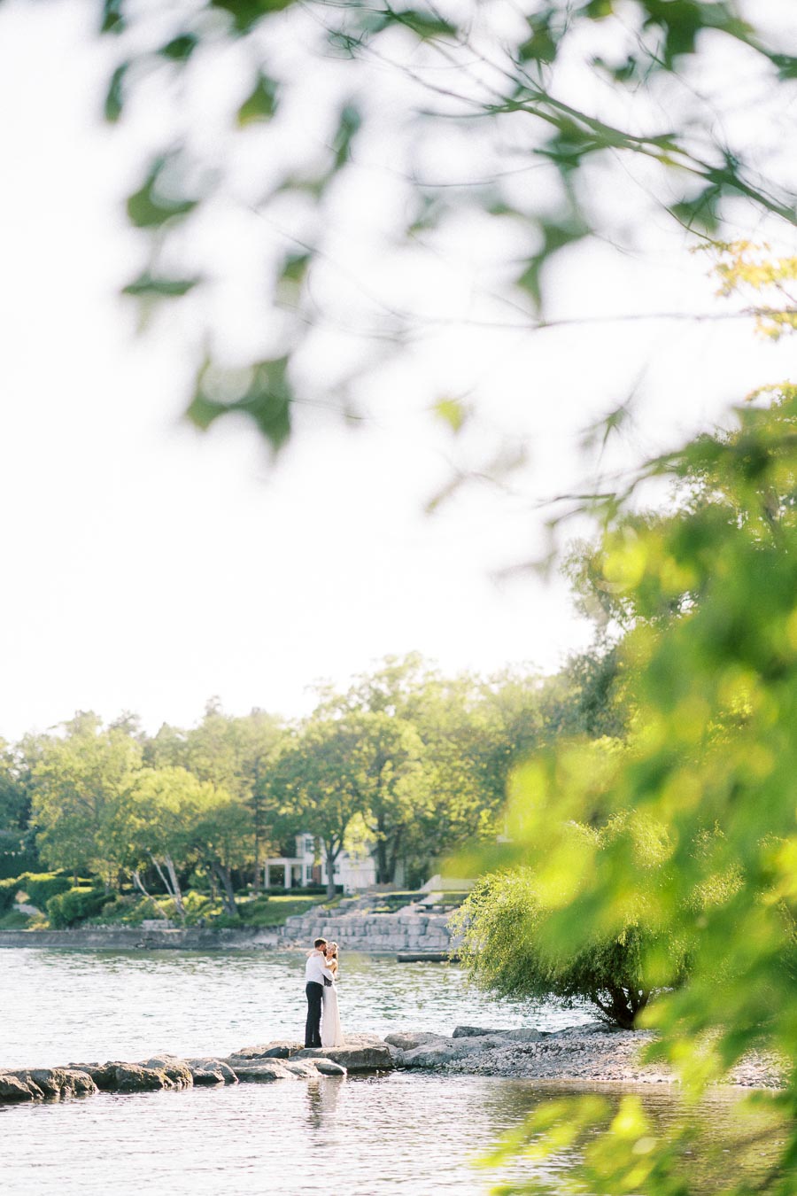 Engaged couple standing on lakeside rocks, embracing with trees and water in background.