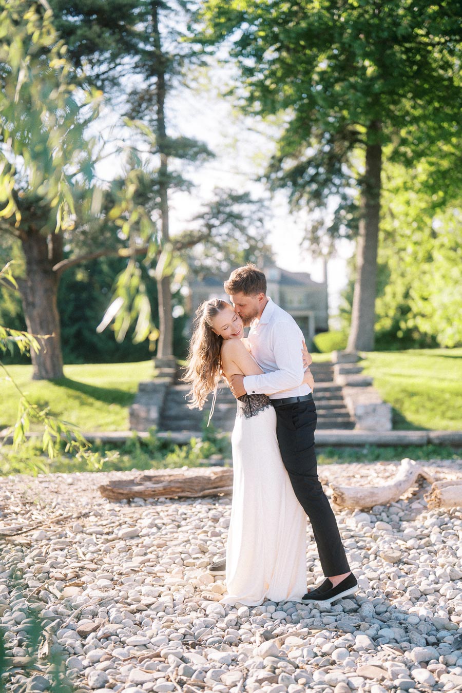 Anastasiia and Petr hugging on a sunlit rocky shore with stone steps and trees behind them.