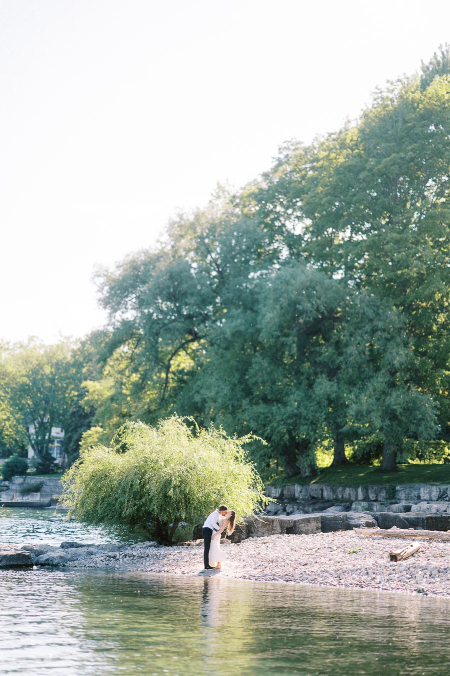 Engaged couple kissing on a rocky shoreline with trees and water nearby.