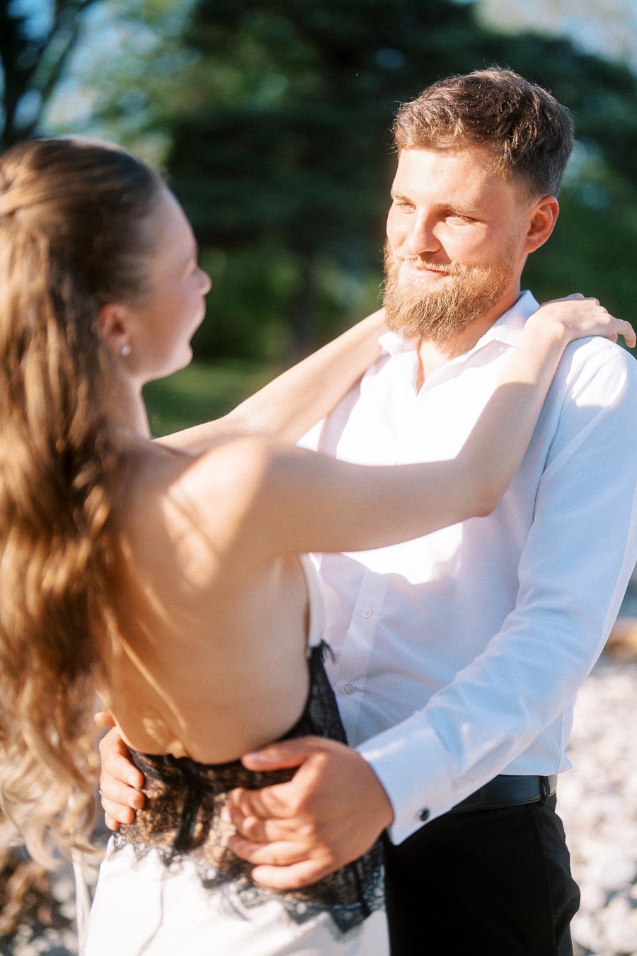 Engaged couple standing outdoors in sunlight, arms around each other, greenery in background.