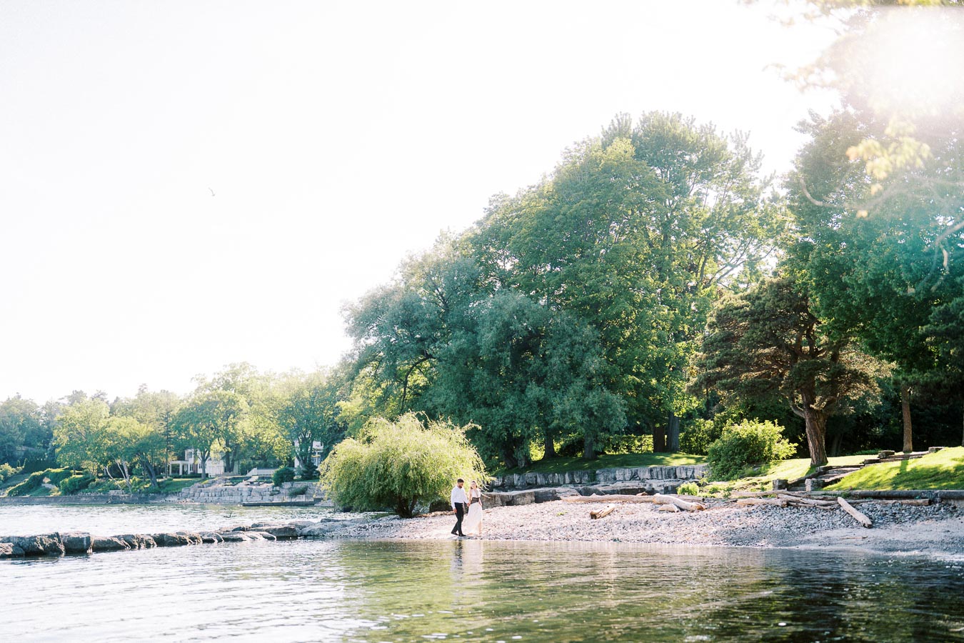 Engaged couple walking together on a sunny shoreline with trees and clear water nearby.