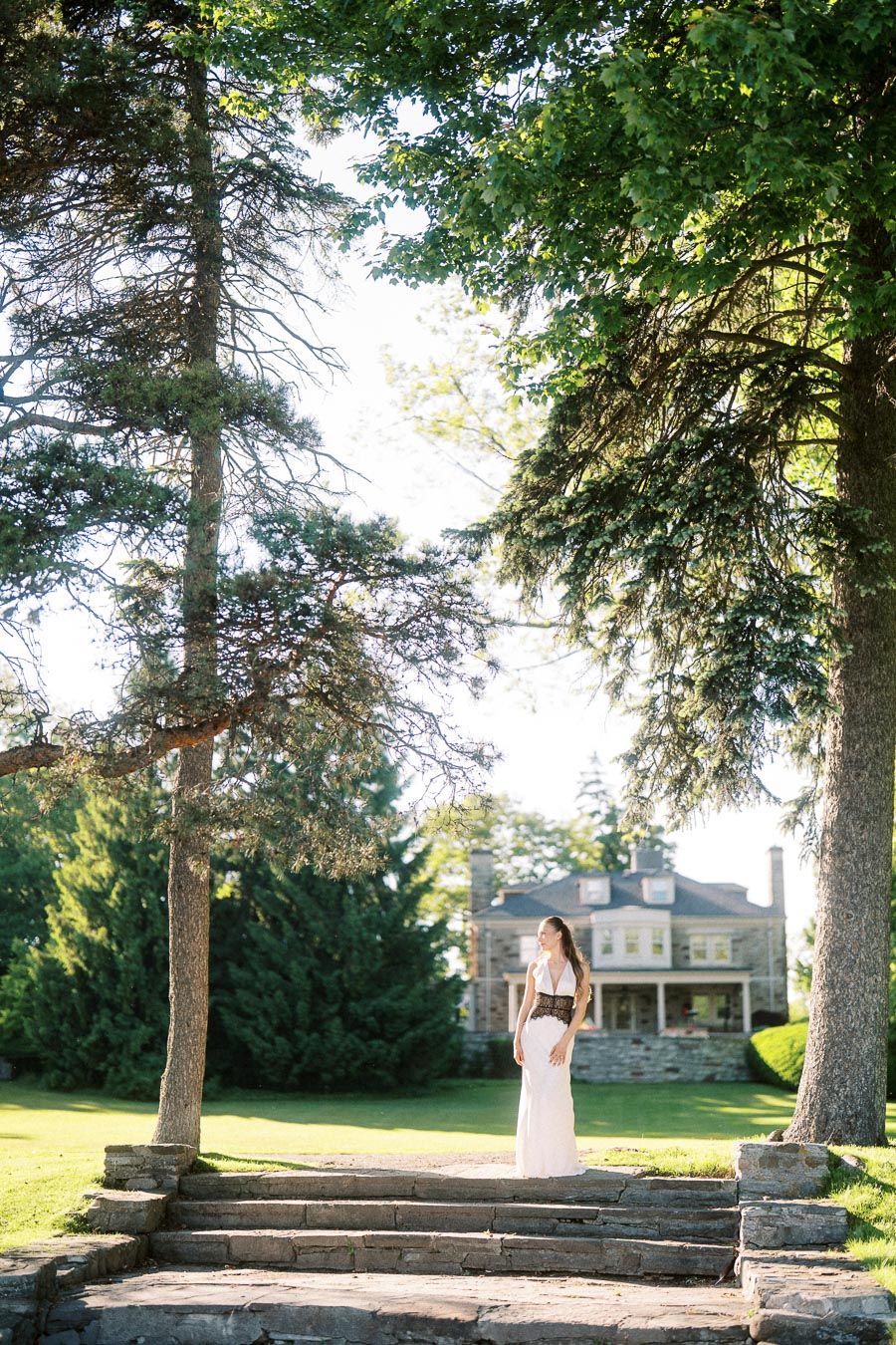 Woman in white dress stands on stone steps beneath large trees, mansion in background.