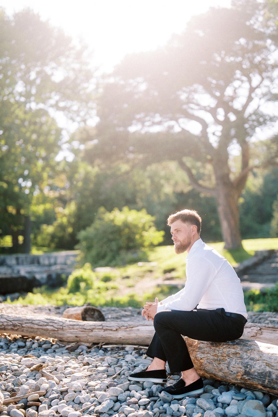 Man in white shirt and dark pants sits on driftwood log beside rocky lakeshore with trees in background.