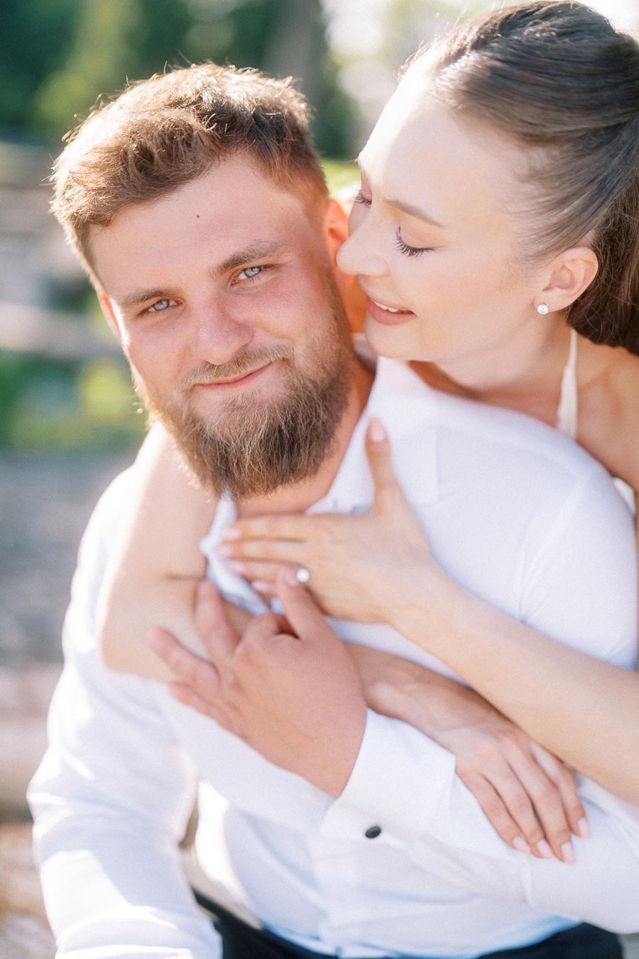 Anastasiia embraces Petr from behind, her engagement ring visible on his chest, both smiling outdoors.