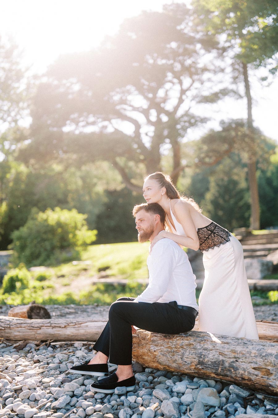 Anastasiia and Petr pose together on a driftwood log at a sunlit, rocky lakeshore with trees in the background.