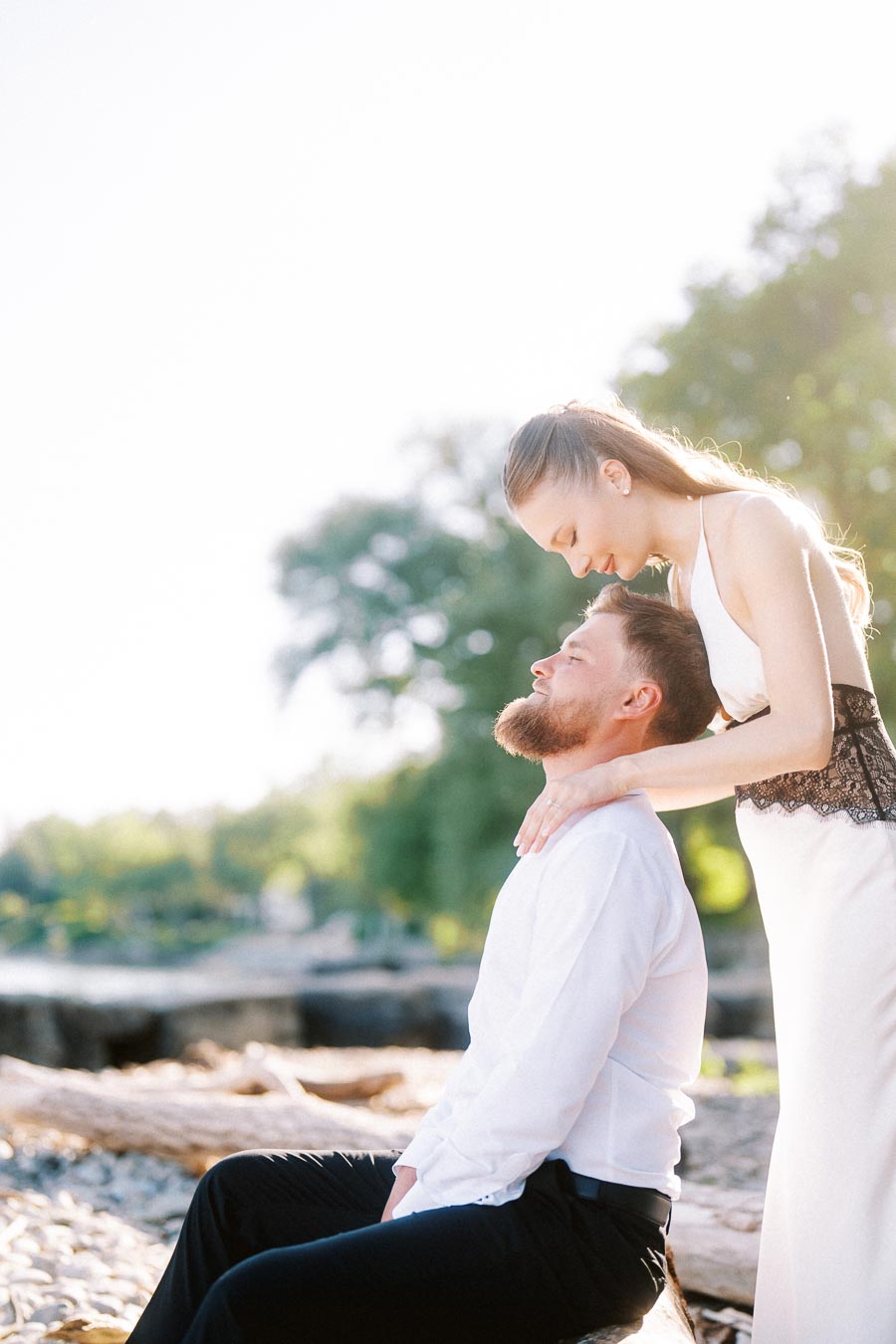 Anastasiia leans forward to touch foreheads with Petr, who is seated on driftwood by the lake.