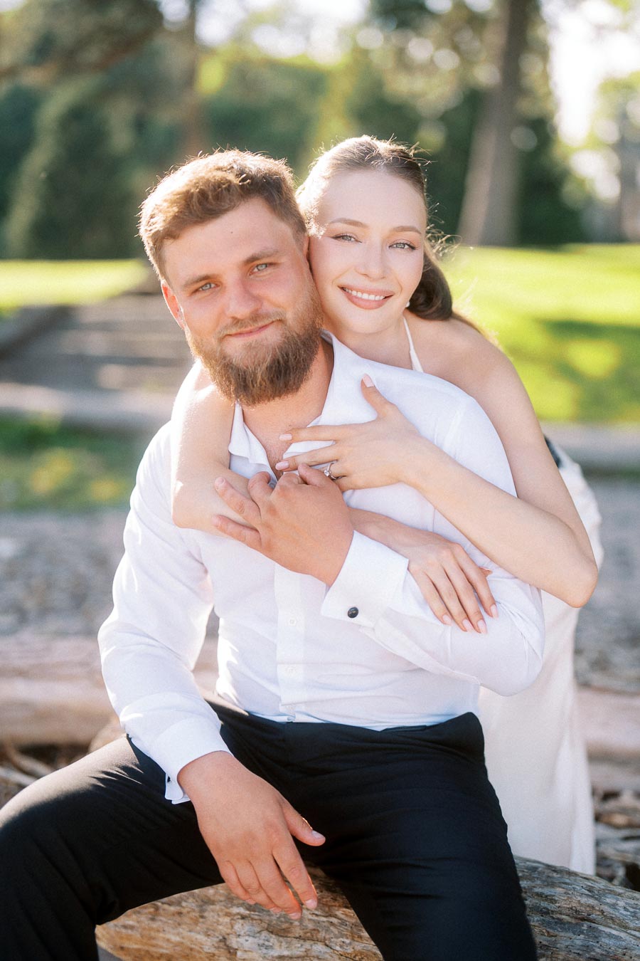 Anastasiia hugs Petr from behind on a park bench, her engagement ring visible, both smiling.
