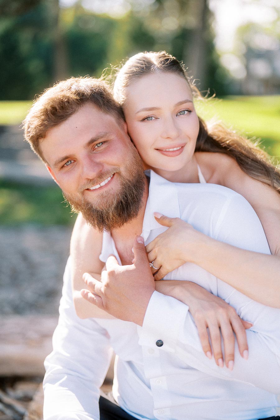 Engaged couple sitting closely outdoors, woman hugging man from behind, both smiling, engagement ring visible.