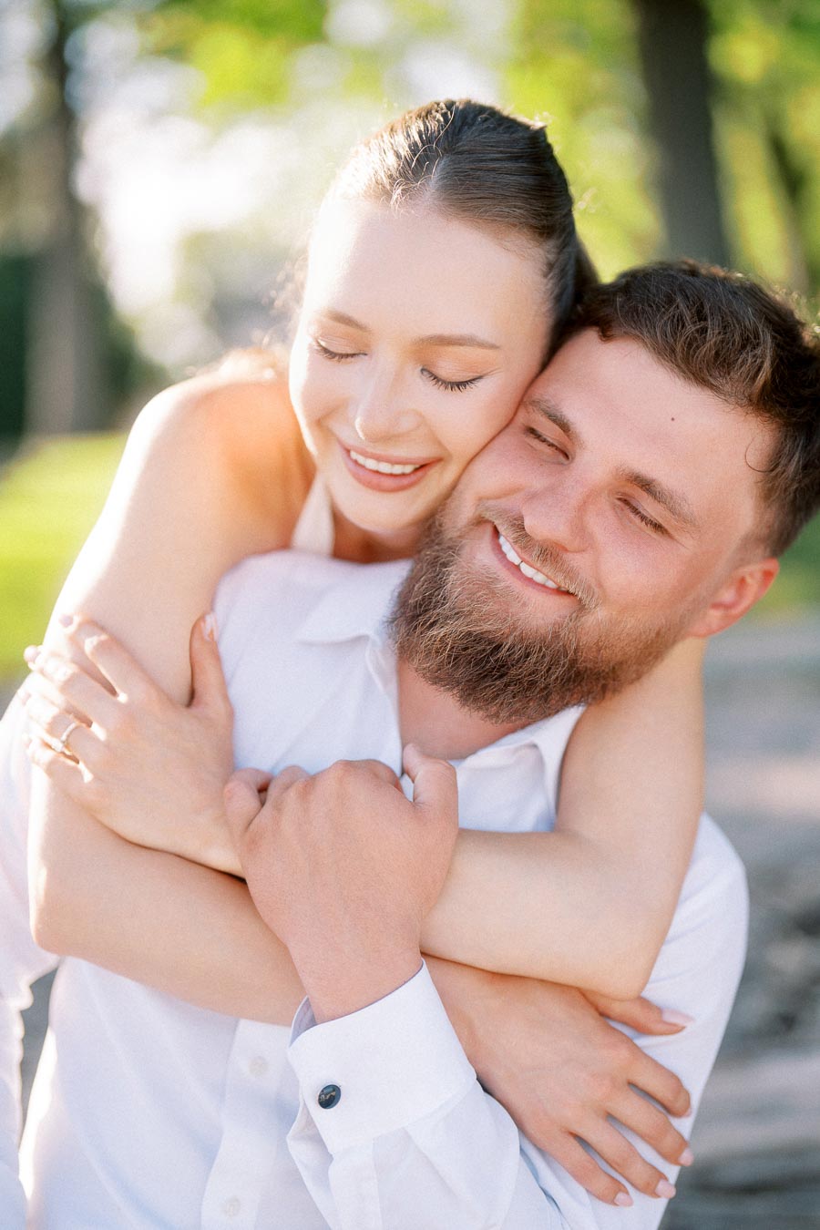 Anastasiia hugs Petr from behind, both smiling with eyes closed in soft sunlight.
