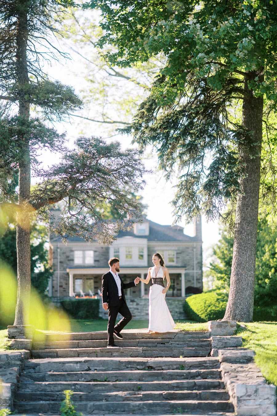 Anastasiia and Petr descend stone steps holding hands, mansion and trees in background.