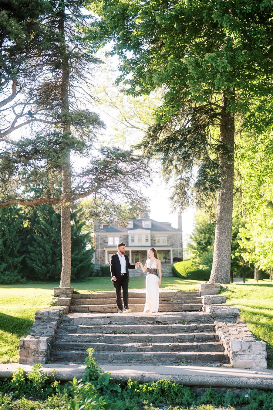 Anastasiia and Petr walk down stone steps beneath trees, Paletta Mansion in background.