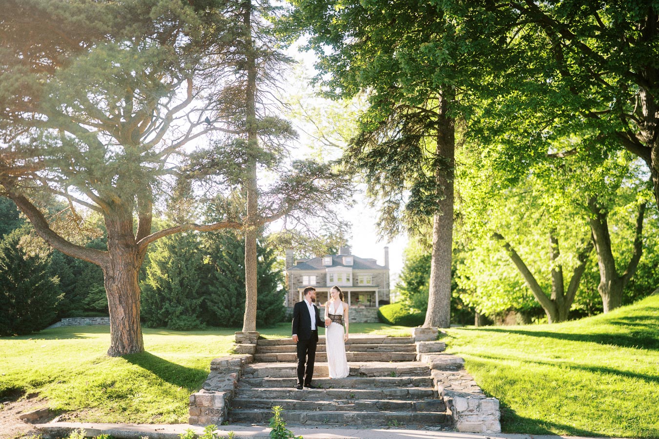 Anastasiia and Petr stand side by side on stone steps, surrounded by trees and greenery, with Paletta Mansion in the background.