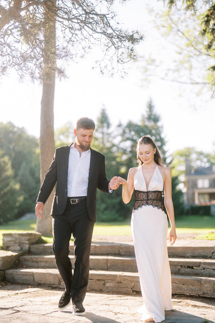 Engaged couple holding hands while descending stone steps in a sunlit park setting.