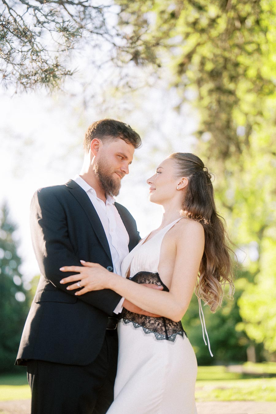 Engaged couple standing closely together beneath trees in soft natural light.