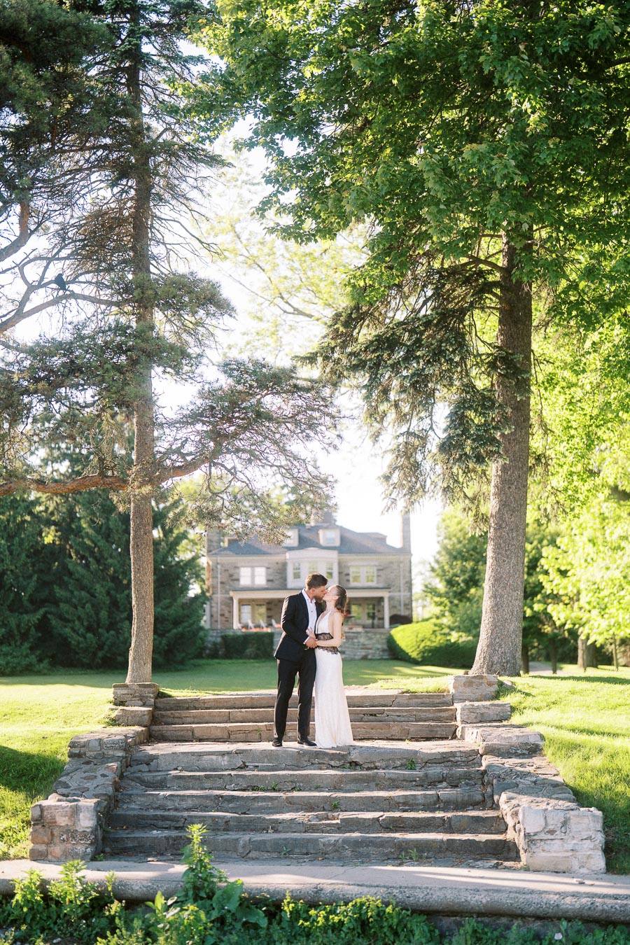 Engaged couple standing on stone steps under trees with mansion in background.
