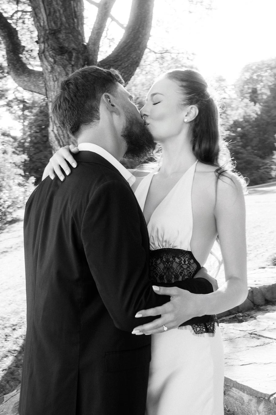 Anastasiia and Petr kissing closely under a large tree in black and white outdoor portrait.