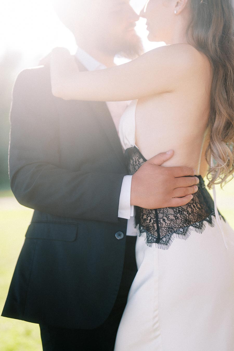 Engaged couple holding each other closely in bright natural sunlight, only torsos and arms visible.