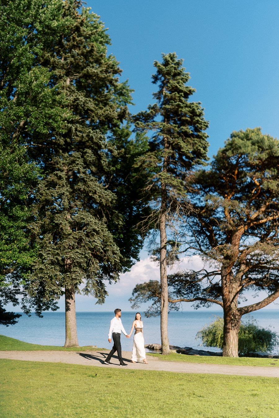 Anastasiia and Petr walk holding hands on a lakeside path bordered by tall trees and grass.