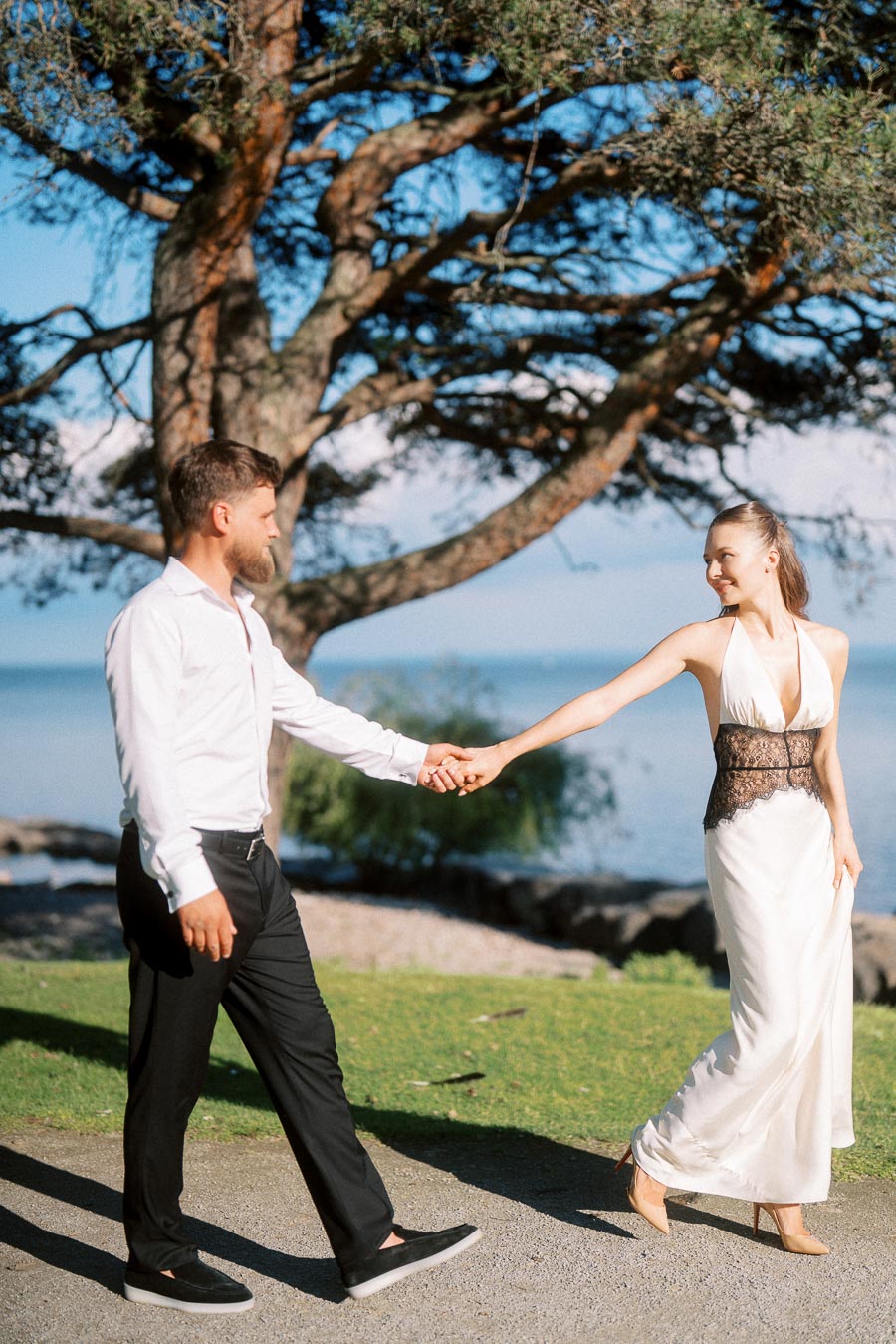 Anastasiia and Petr holding hands and walking near Lake Ontario with a large tree behind them.