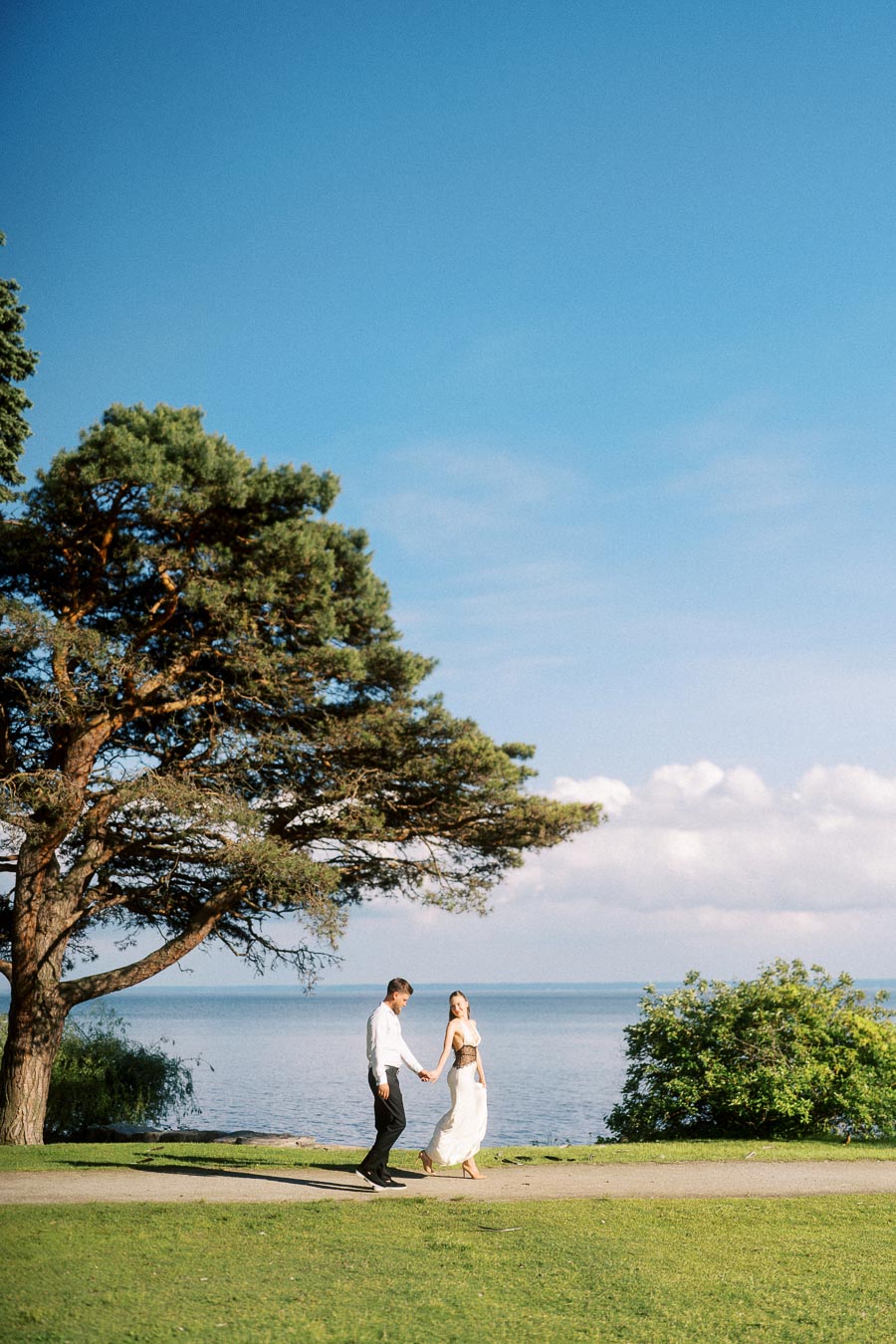 Anastasiia and Petr holding hands while walking on a grassy path beside Lake Ontario with a large tree nearby.