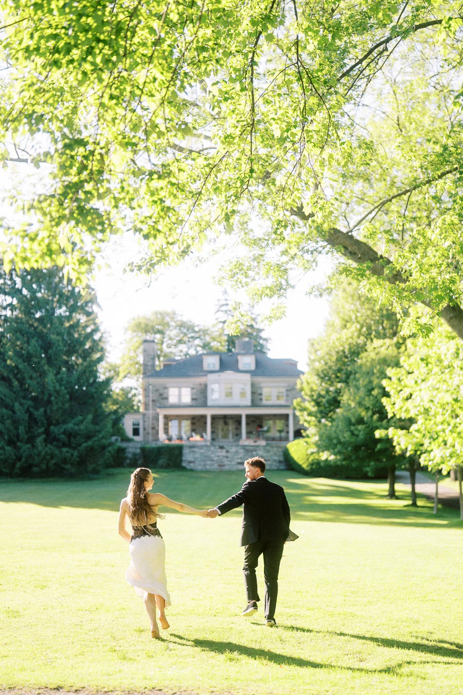 Engaged couple holding hands and walking barefoot on green lawn toward Paletta Mansion under leafy trees.