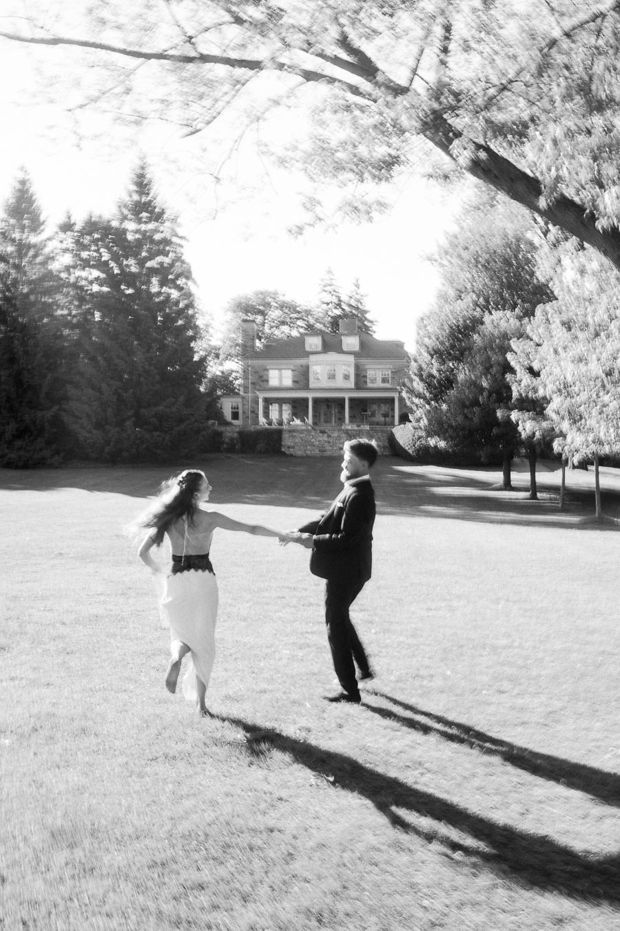 Anastasiia and Petr spinning hand in hand on grassy lawn with Paletta Mansion in background, bright sunlight, black and white.