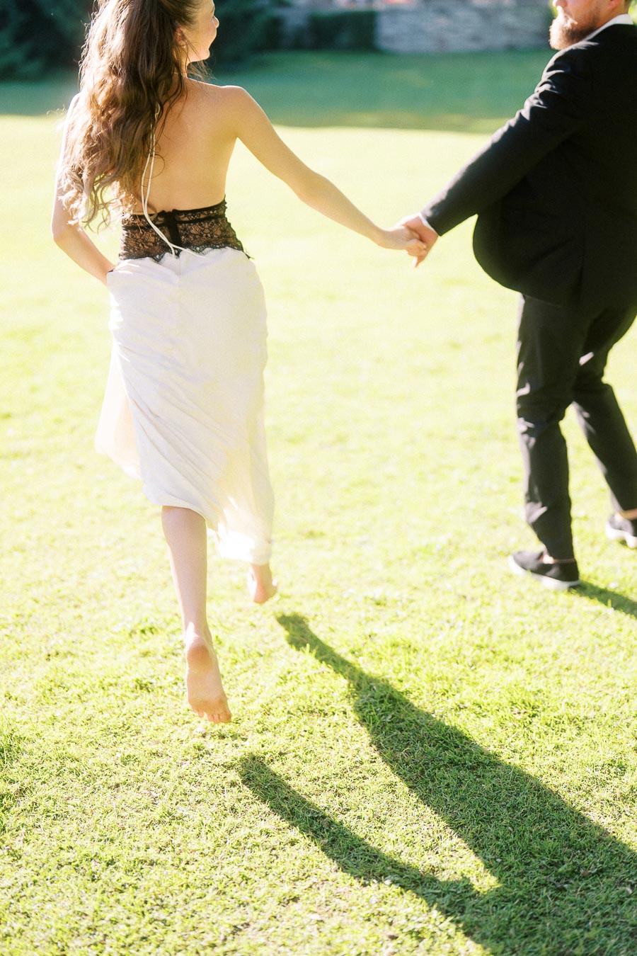 Engaged couple running barefoot on bright green lawn, holding hands, photographed from behind.