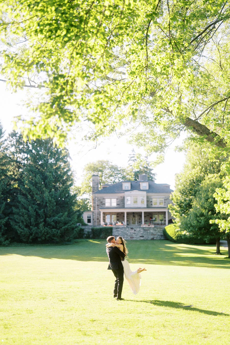 Engaged couple embracing on green lawn with Paletta Mansion in the background under leafy trees.
