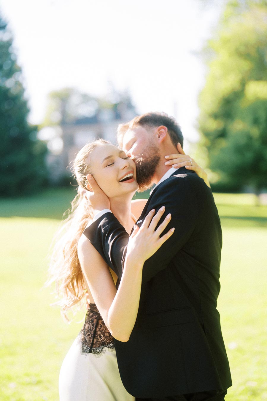 Engaged couple embracing closely in bright sunlight on a green lawn with mansion in background.