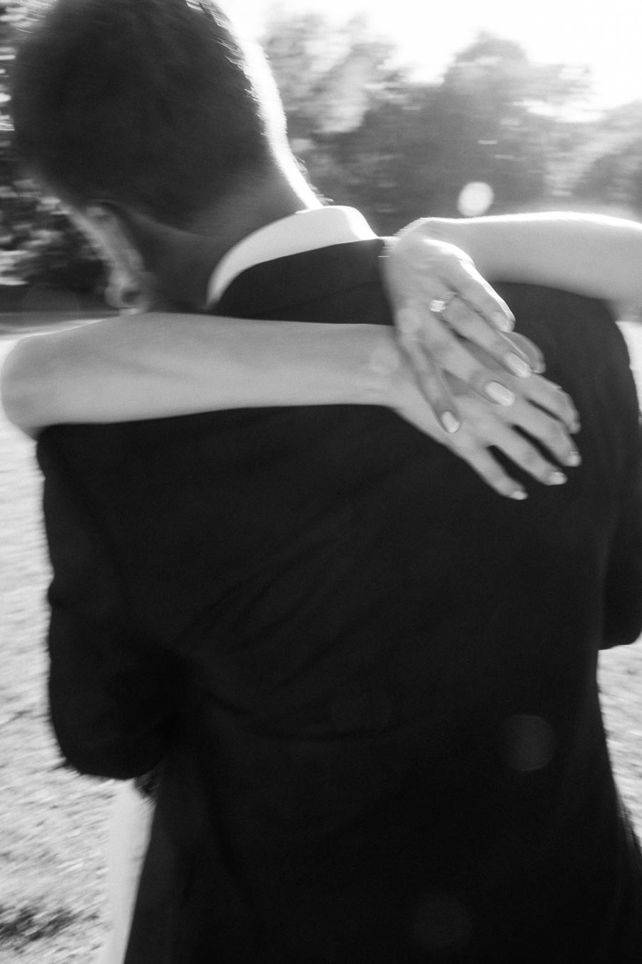 Engaged couple embracing closely outdoors, bride-to-be's hand with ring on groom-to-be's shoulder, black and white photo.