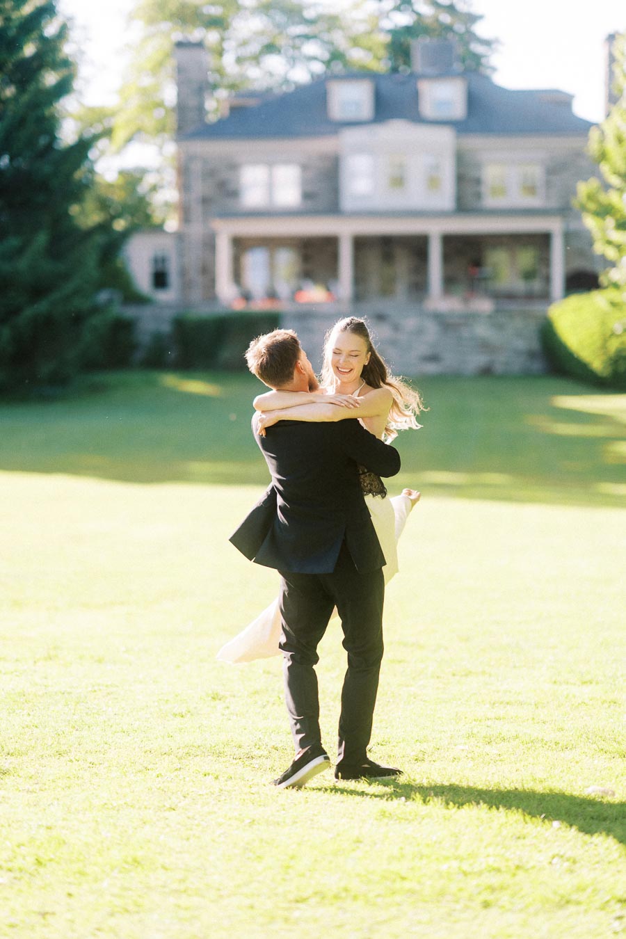 Engaged couple embracing on green lawn with stone mansion in background, bright sunlight.