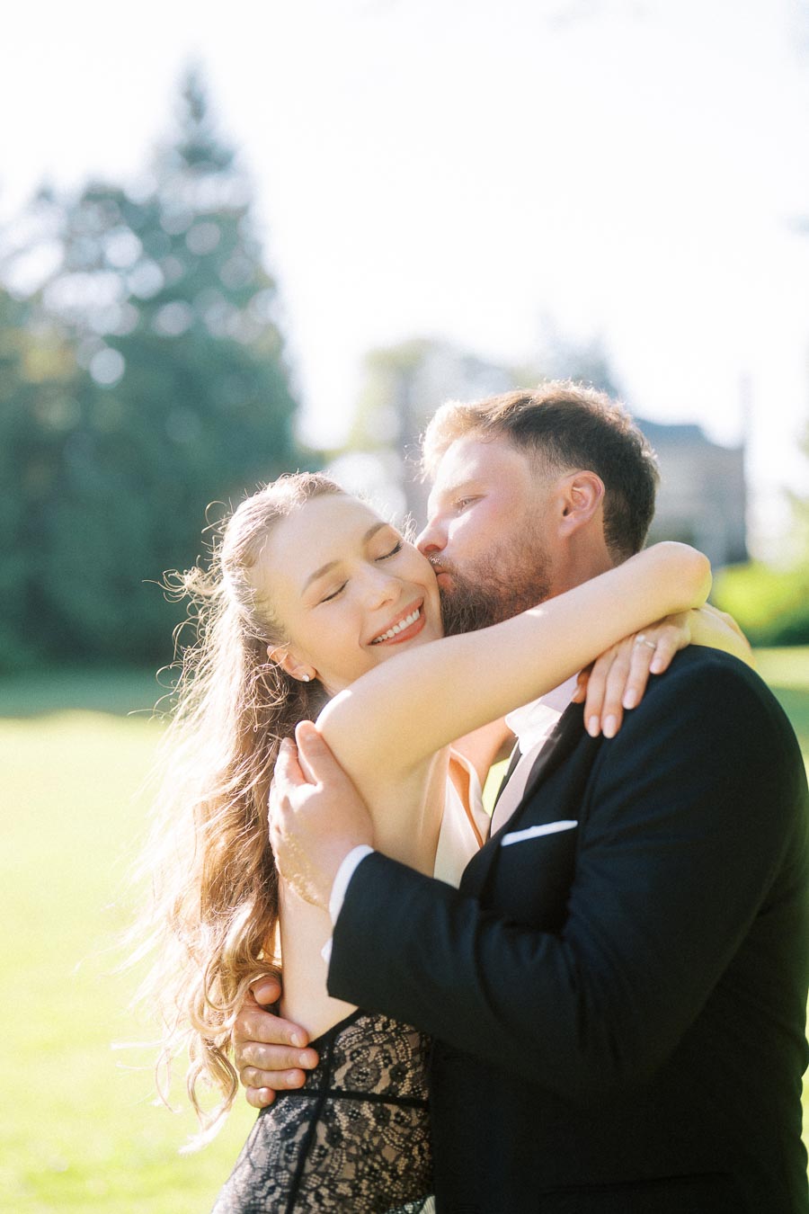 Anastasiia and Petr embracing outdoors, Petr kissing Anastasiia's cheek, sunlit green lawn behind them.