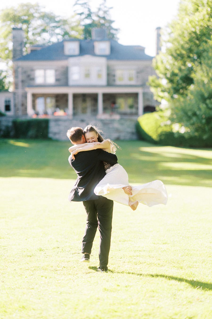 Engaged couple on green lawn, Petr lifting Anastasiia, Paletta Mansion in background, bright sunlight.