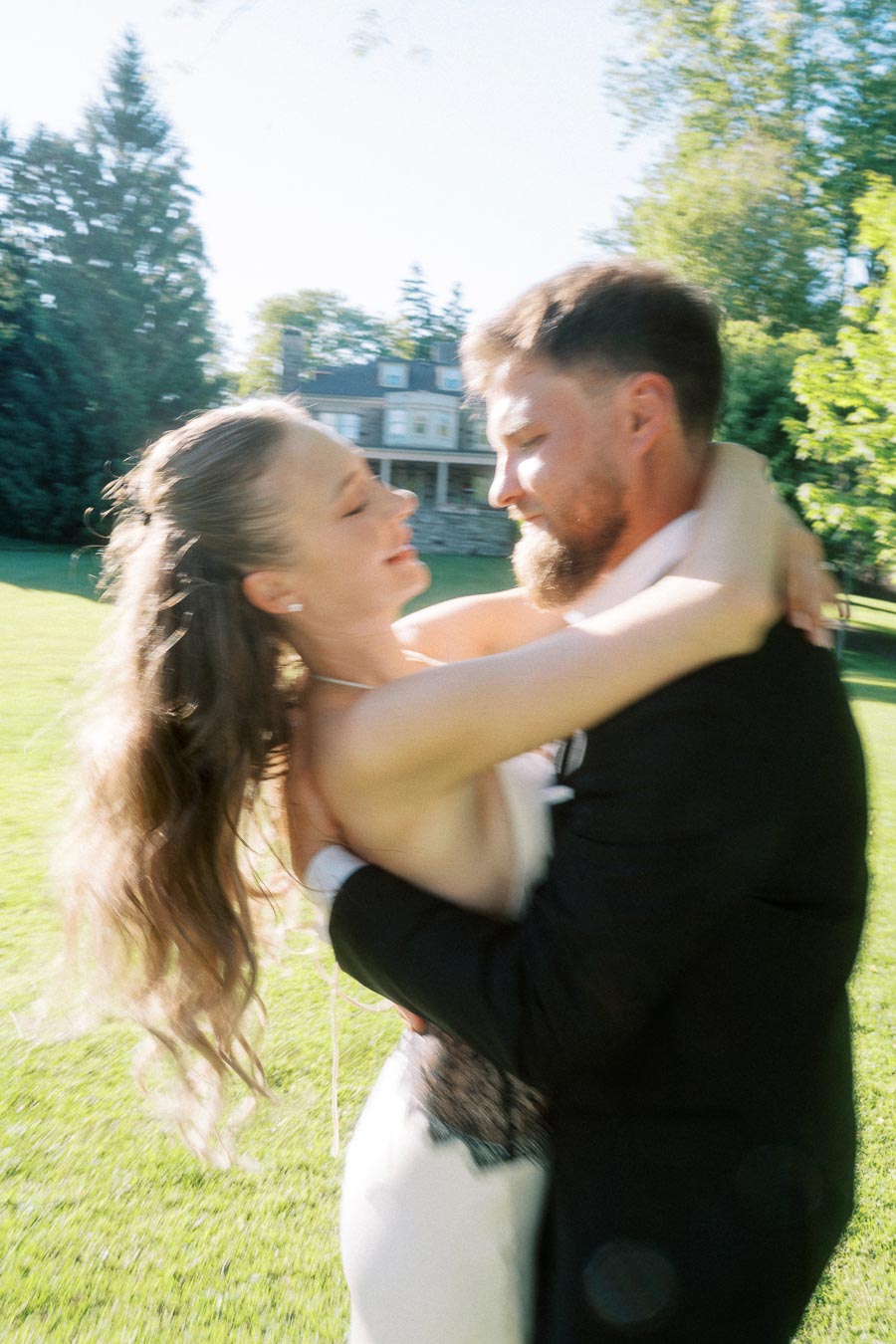 Engaged couple embracing on green lawn with mansion and trees behind them.