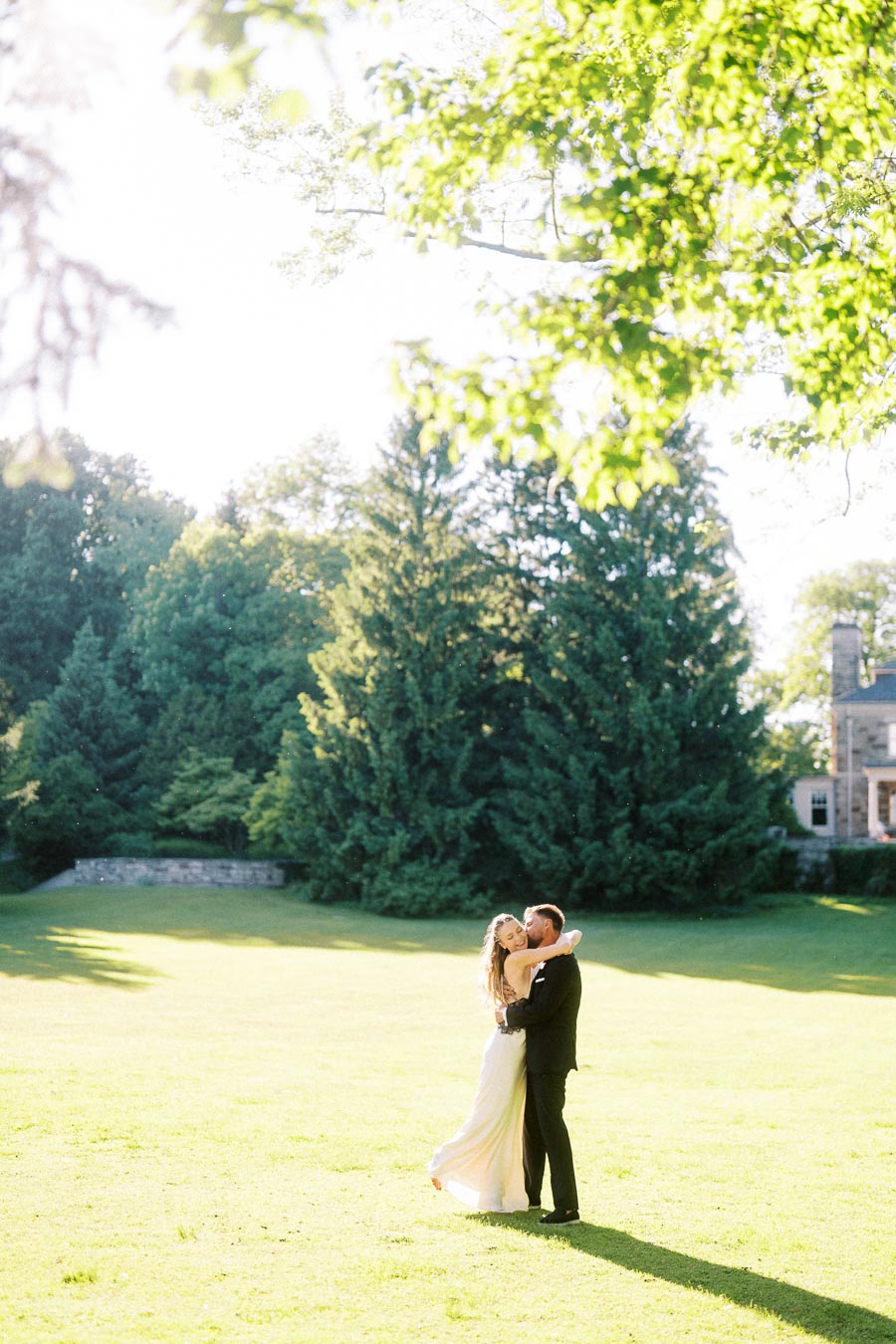 Engaged couple embracing on a sunlit lawn with trees and stone building in background.