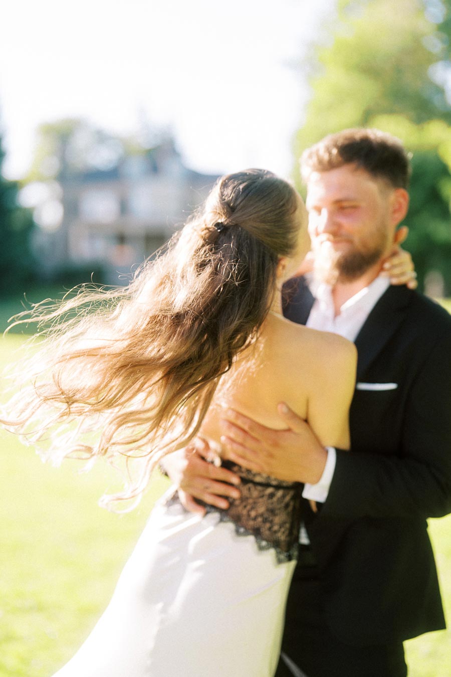 Anastasiia and Petr hugging on a grassy lawn with sunlight and blurred mansion in background.