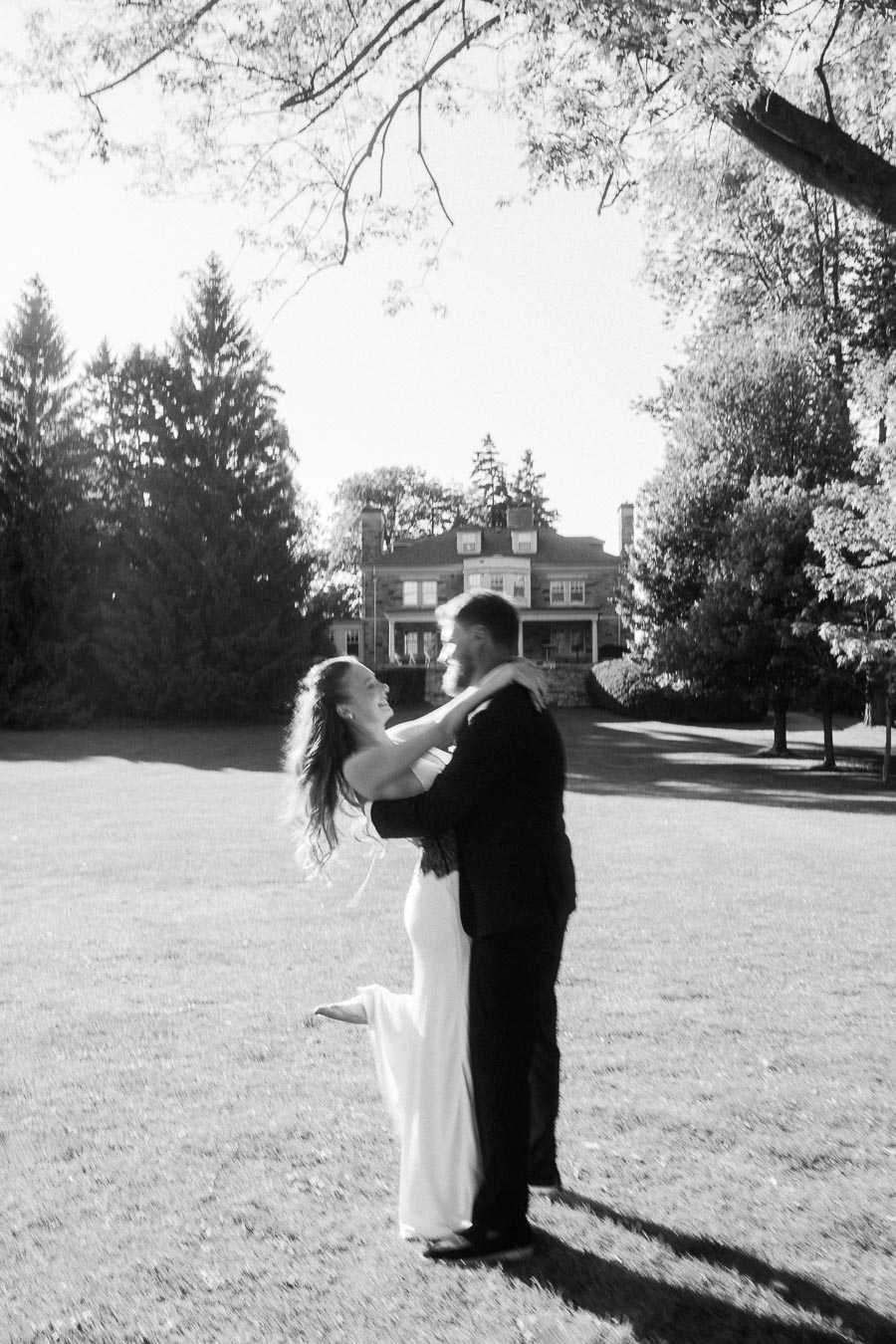 Anastasiia and Petr spinning together on a sunlit lawn with Paletta Mansion in the background, black and white photo.