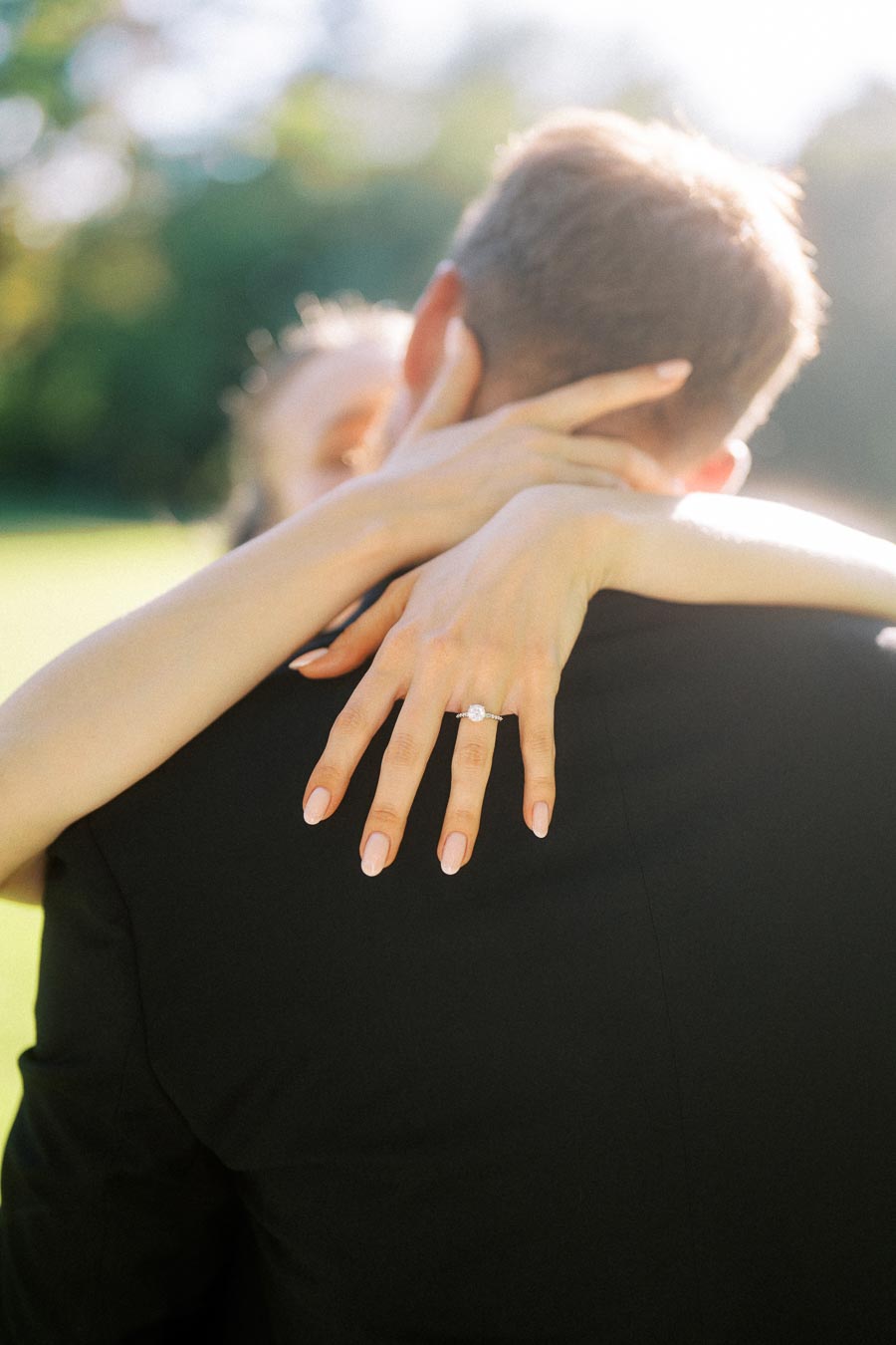 Close-up of Anastasiia's hand with engagement ring resting on Petr's shoulder during an embrace outdoors.
